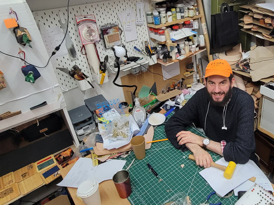 Shows the inside of a creative workshop with a bearded man with an Orange hat sitting at his workbench with a yellow mallet. Surrounded by printers, hand tools and paints.