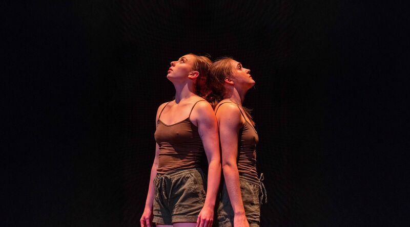 Two dancers stand back to back, looking up.