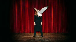A woman is stood on a theatre stage in front of a red curtain. She’s white, wearing all black and has long brown hair. A seagull is flying towards her, blocking us from seeing her face.  