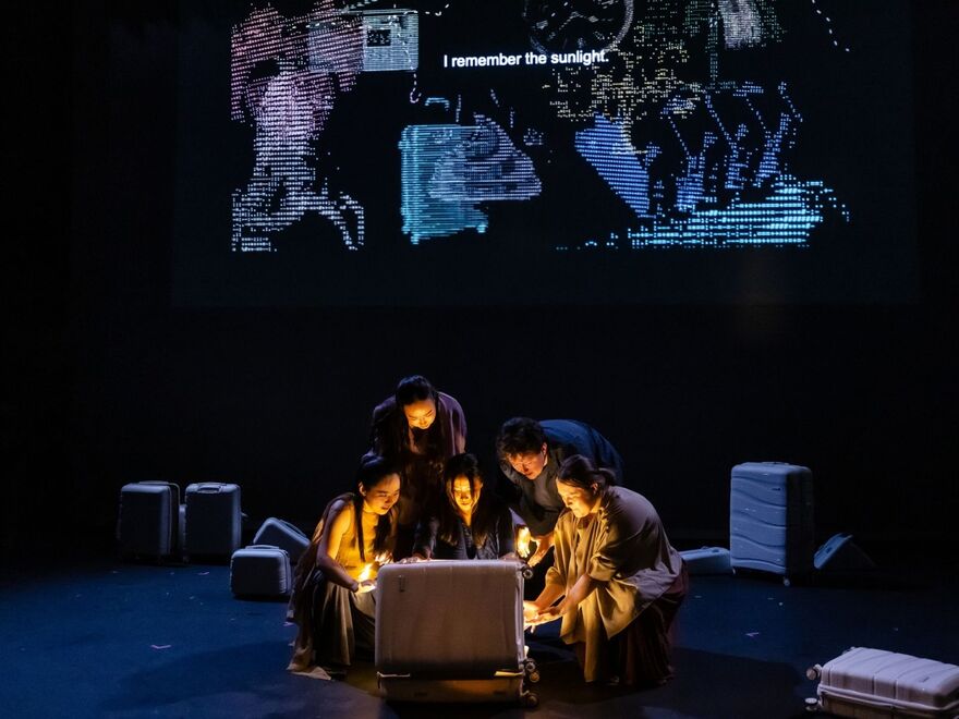 An image of five actors crowded around an open suitcase which shines a warm orange light in their faces. Above them, you can see the pixelated outlines of various images projected above their heads. 