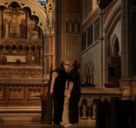 Two people wearing black, standing close to each other looking up. Neck exposed. In a church.