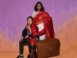 Two Performers One Is Sat By A Suitcase The Other Is Stood Behind With Hand On Shoulder