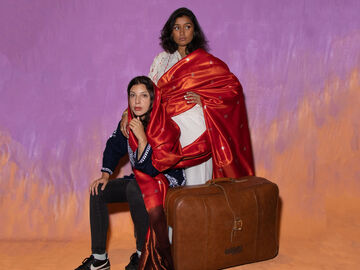 Two Performers One Is Sat By A Suitcase The Other Is Stood Behind With Hand On Shoulder