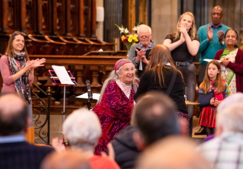A Smiling Person With Grey Hair In A Pink-Red Dress And red headband Is At The Front Of A Crowd Shaking Hands
