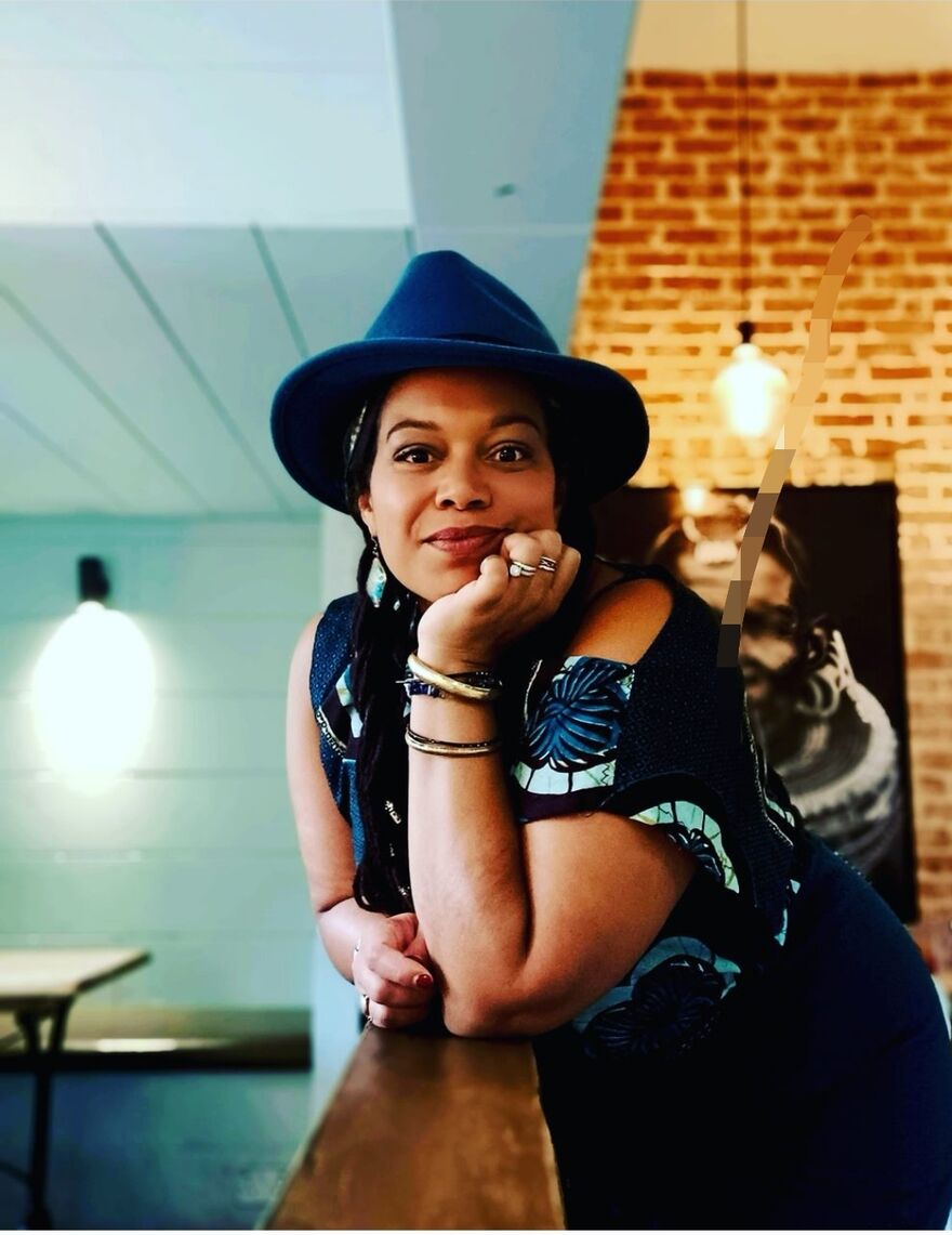 Fée Uhssi is a women of  Franco-Nigerian background in her 40s. She is wearing one of creation, a block top in a teal print and a matching skirt and hat. 
