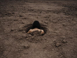 A young woman is submerged in a hole in an empty field of dirt. Only the back of their head and shoulders is visible. 