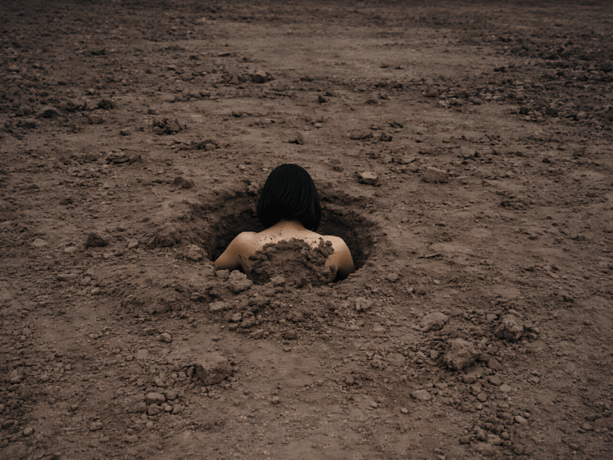 A young woman is submerged in a hole in an empty field of dirt. Only the back of their head and shoulders is visible. 
