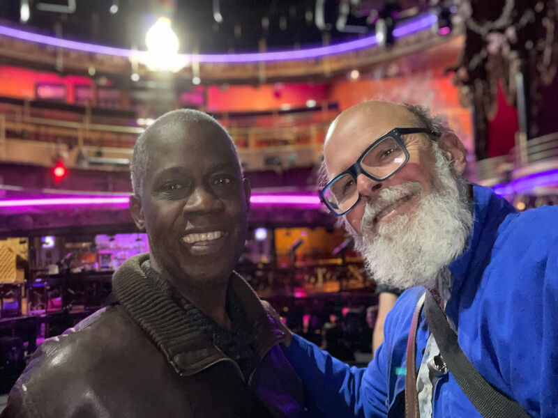 Two, older men standing together on the stage facing the camera at the Clapham Grand, with the auditorium behind them. The man on the left has a white complexion and a full, white beard. The man on the right has a black complexion and a shaved head. Both are smiling.
