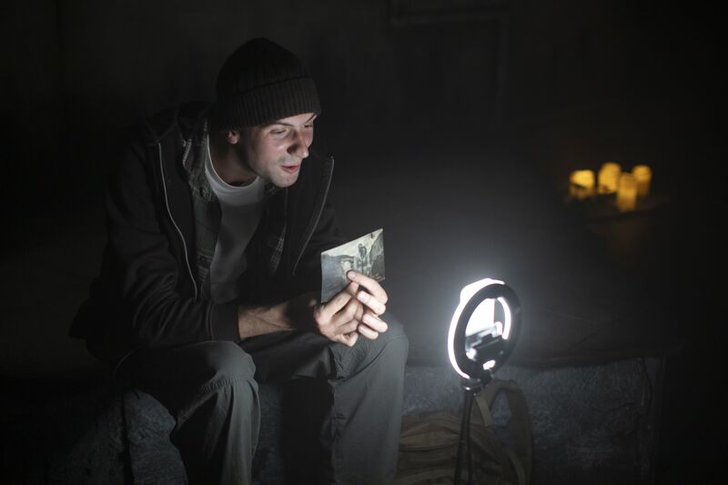 A man in a black jacket, trousers and beanie hat is holding a black and white photo. He is using a self-ring that is glowing in front of him.