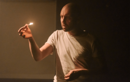A bald man in a dimly lit room with white walls is wearing a white t-shirt and holding a lit match aloft, staring at it seriously.  