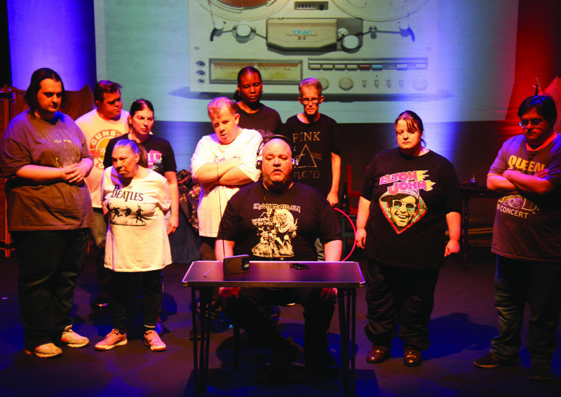A group of people stand around a man who is sat at a table centre stage and is wearing headphones. Everyone looks sad or concerned and they are staring at the man in the middle. The man has a stressed expression on his face. The group are all wearing band t-shirts and there is an image of a tape recorded projected behind them. 