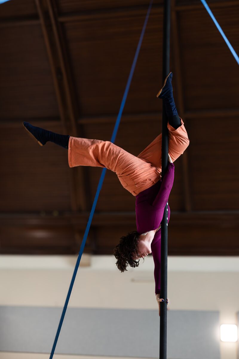 A person is upside down on a vertical pole, with one leg out behind them