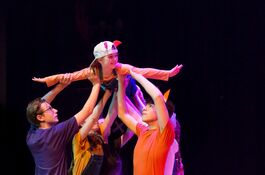Four young people hold up another young person in the sky. They are dressed up as foxes and the young person in the sky has their arms stretched out like they are flying.