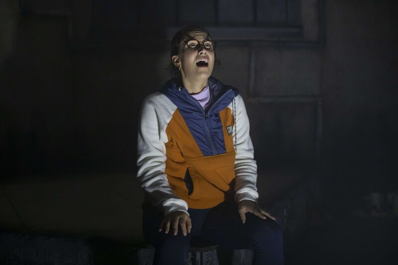 A woman in an orange and white jacket is sat down with her hands on her knees. She is screaming.