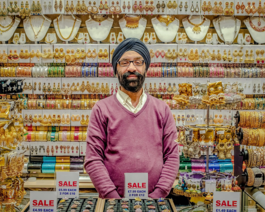 Image shows Kirpal Nagpal behind the counter of his shop, KNG designs in Tooting. Behind him and in front of him you can see brightly coloured gold costume jewellery.
