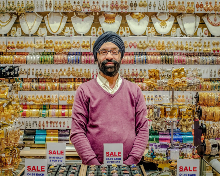 Image shows Kirpal Nagpal behind the counter of his shop, KNG designs in Tooting. Behind him and in front of him you can see brightly coloured gold costume jewellery.