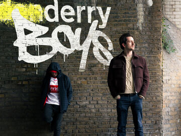 Two men stood in front of a wall where the words Derry Boys are spray painted on