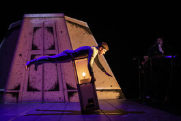 A male performer dressed in a blue onesie is balancing on a light as it tips over, with his legs and arms in the air. 