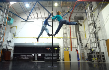 Two women climb a pole, facing each other. They each have one foot on the pole with their other leg extended away in the air. They are 2m from the floor.