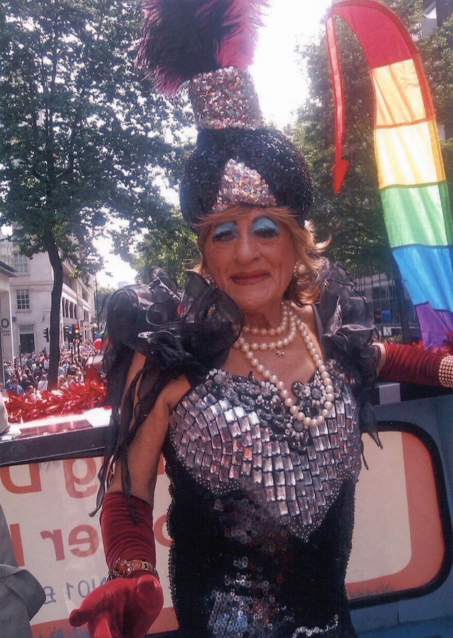 Person in a Pride parade in drag wearing a sequinned black and silver outfit, tall feathered headdress, pearl necklace and red gloves, standing on a float with a rainbow flag visible behind.