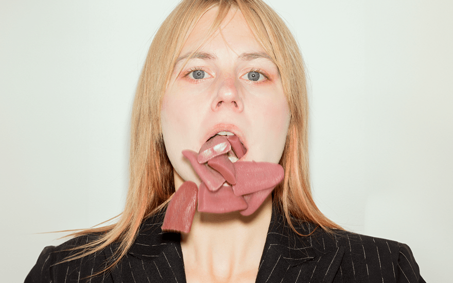 A headshot of a white woman with blonde hair and blue eyes in a black and white pinstriped suit. Her mouth is open, and lots of tongues are falling out of it.