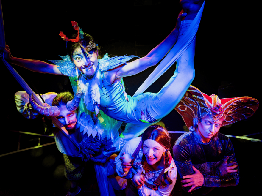 Four performers in elaborate costumes, with one suspended in mid-air on aerial silks. The others pose below in dramatic outfits against a dark background.
