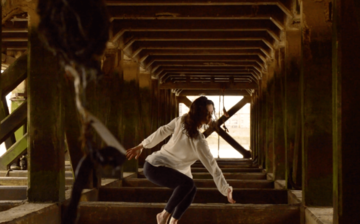 Woman dances under Pier along the River Thames, wearing white shirt and black trousers with hair and clothes wet.