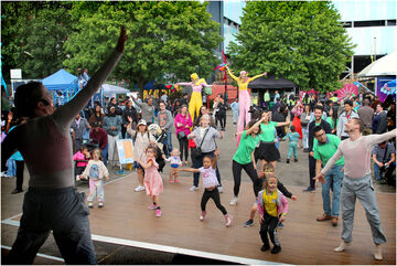 Adults and Children at a festival outdoors dancing in front of a stage, with stilt walkers in the background