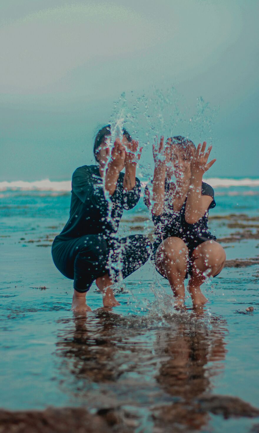 Two people splash water at the shoreline, droplets rising against the sea and sky.