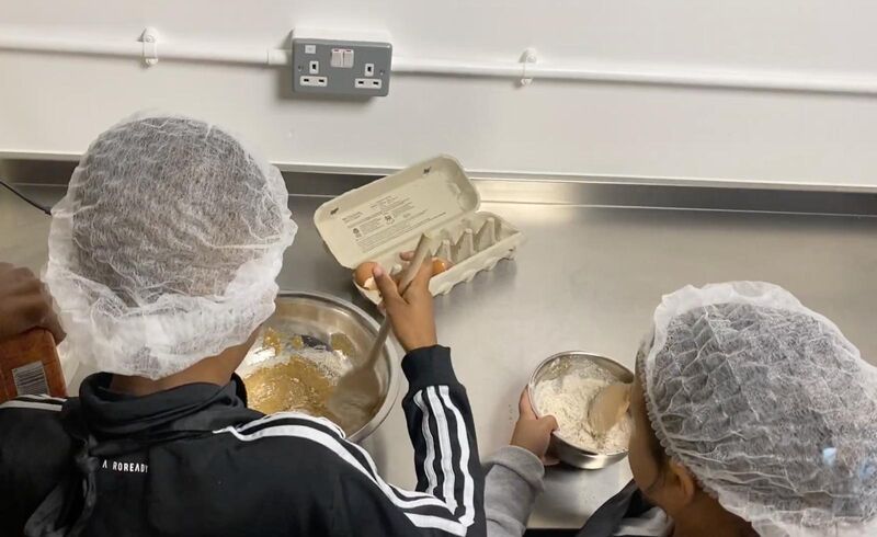 Two young children crack eggs and create a batter mix in a stainless steel bowl. The bowl sits on a stainless steel kitchen surface.