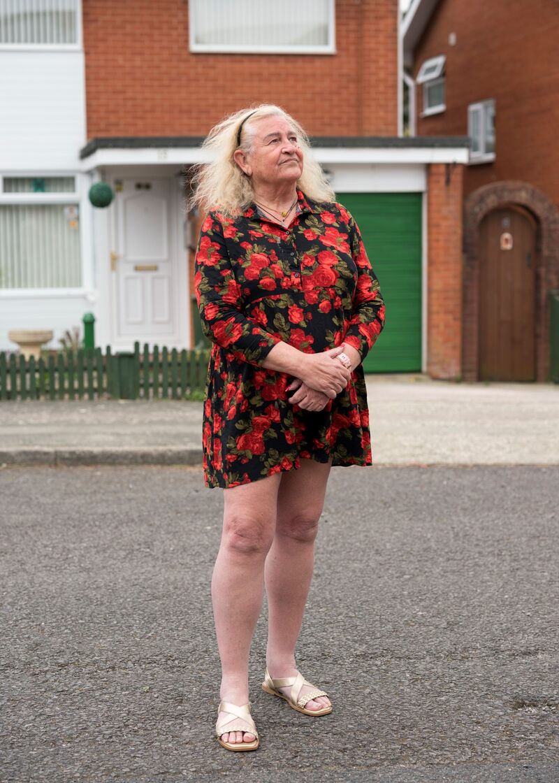 Photographic portrait of Allana, a white woman in her seventies who is wearing a short dress with red flowers on it. She has blond hair and is clasping her hands in front of her.