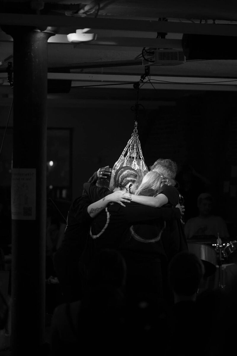 black and white image of members of the Wild Mix choir embracing around a transparent kickboxing bag instrument