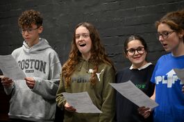 4 young people stand holding white paper. They are smiling and have their mouths open as they are singing.