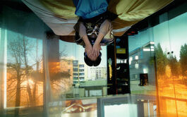 An image of a young person sitting on the edge of their bed with their hands covering their face. The young person and the bed are situated upside down as if stuck to the ceiling. The walls around the room have projections of buildings, trees and outdoor spaces. 