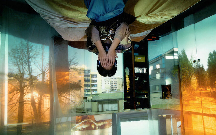 An image of a young person sitting on the edge of their bed with their hands covering their face. The young person and the bed are situated upside down as if stuck to the ceiling. The walls around the room have projections of buildings, trees and outdoor spaces. 