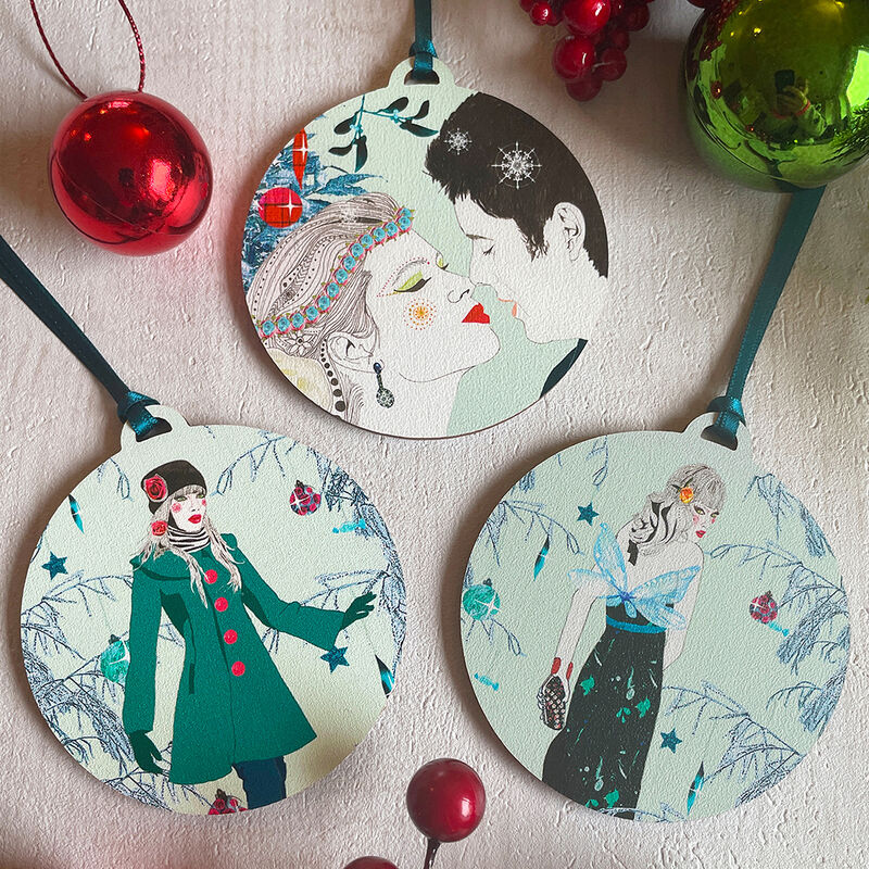 A set of three, round, birchwood Christmas Decorations with teal ribbons. Designs from left to right: Ice Skating Maiden, Christmas Mistletoe Kissing and Magical Christmas Fairy.