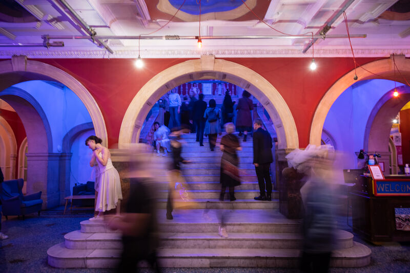 People stood on stairs underneath some archways, some figures are in motion