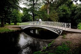 A decorative white iron footbridge arches over the River Wandle in a lush, green park. Two people walk across the bridge, surrounded by tall trees and dense foliage reflected in the calm water below.