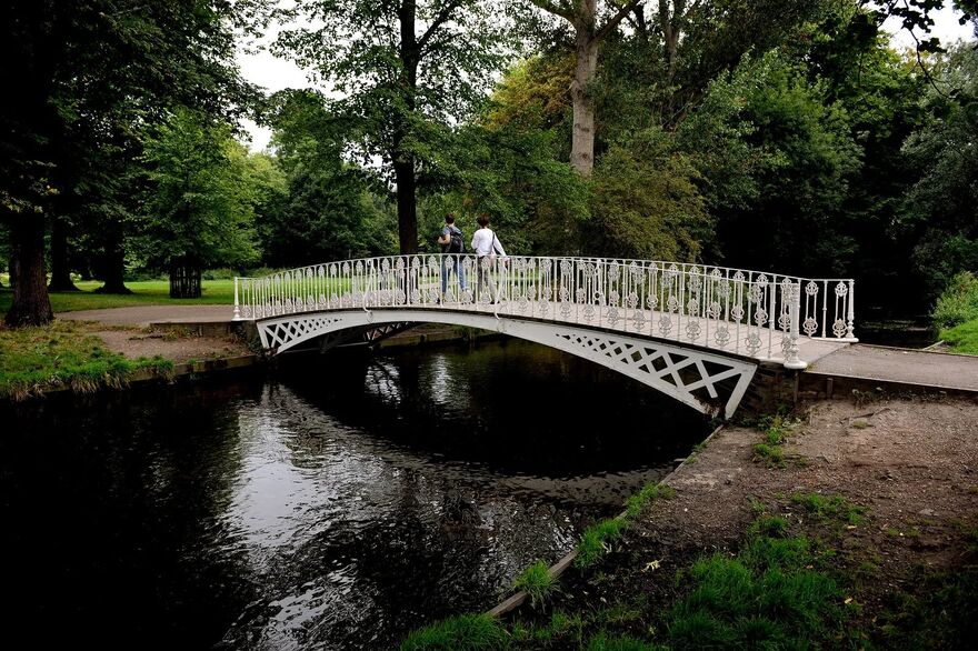 A decorative white iron footbridge arches over the River Wandle in a lush, green park. Two people walk across the bridge, surrounded by tall trees and dense foliage reflected in the calm water below.