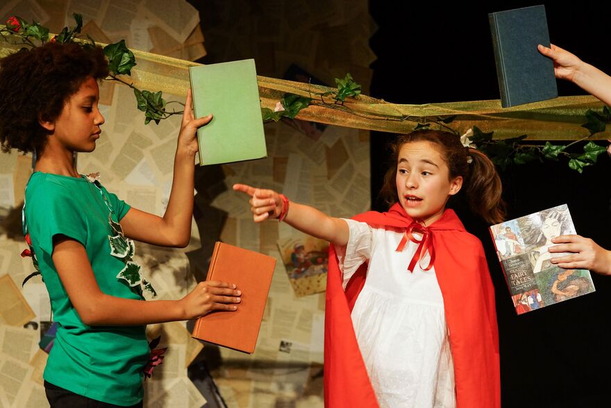 A young girl with brown hair in pigtails points to the lefthand corner of the photograph. There are two books floating by hear head held by more young people out of the shot. 