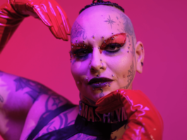 Smashlyn Monroe portrait, with their hands framing their face in red PVC gloves with a hot pink background