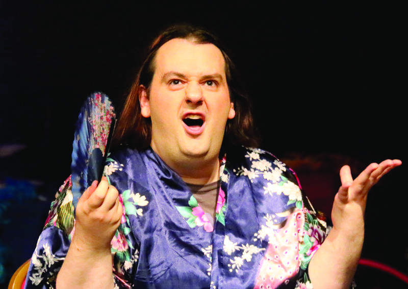 A woman sat with a dramatic expression on her face. She is mid rant, and has a shocked and disgruntled look on her face. She is holding a fan in one hand and wafting it. She has a brown bob hair cut and is wearing a blue silk kimono-type dressing gown with floral designs.