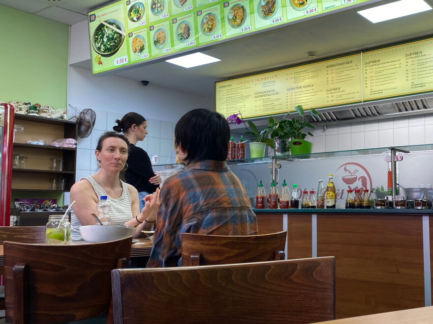 Interior of a cafe. There are people seated at a table facing each other engaged in conversation with the remains of their lunch on the table.