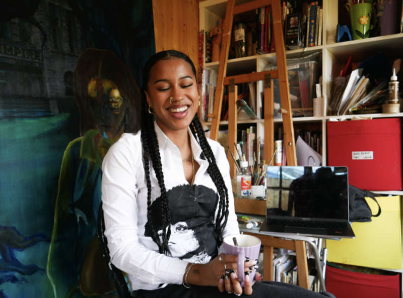 A smiling young woman sat in her studio with a painting and paints behind her