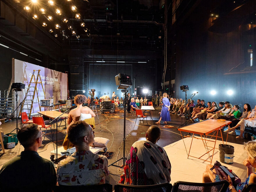 Theatre studio space with audience seated around the edge of the space. There are performers across the stage standing in various positions. There tables, chairs, theatre lamps and a projection screen spread across the space.