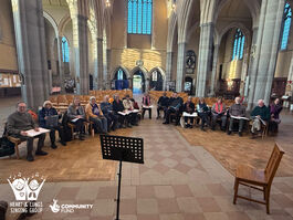 The friendly and diverse members of Heart & Lungs Singing Group, seated, smiling at the camera and ready to begin their singing session in their beautiful venue of All Saints Church, Tooting. 