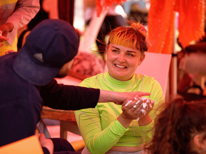 Vicki, a white woman with brightly-coloured hair and wearing a high-vis top, is playing with shaving foam. A workshop participant reaches out to feel the foam.