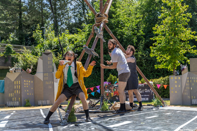 A female performer in a bright yellow raincoat is frantically eating a sandwich while two other performers operate pigeon puppets, hungering for the same food.